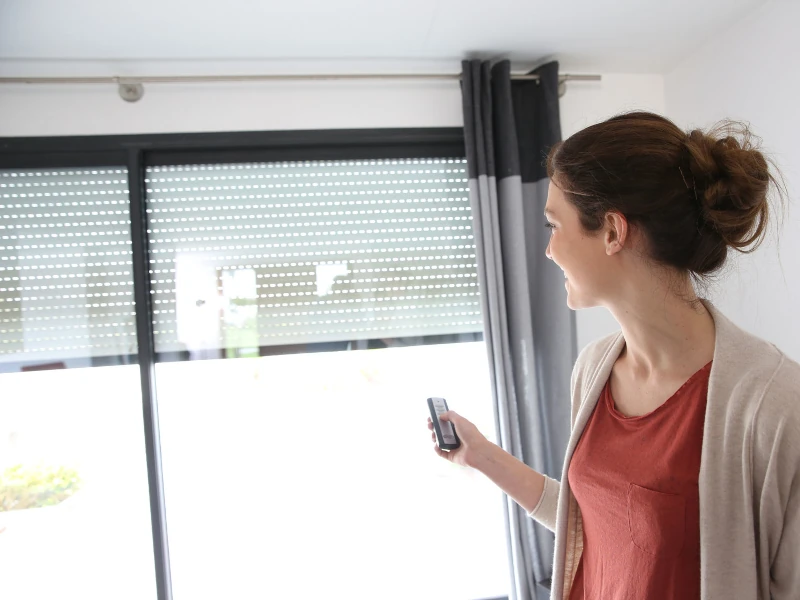 Frau steuert Rollladen per Fernbedienung am Fenster, Smart Home Sonnenschutz